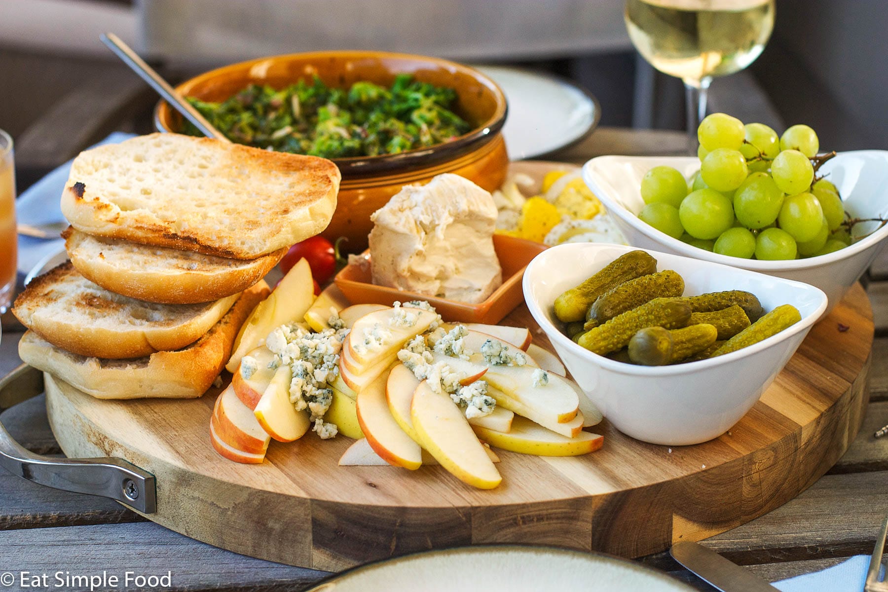 Multiple bowls of food on a wood cutting board. brie, apples with blue cheese, cornichons, green grapes, slices of ciabatta, green salad.