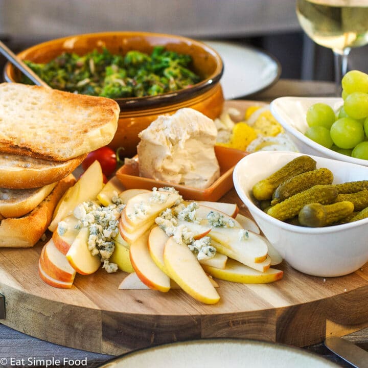 Multiple bowls of food on a wood cutting board. brie, apples with blue cheese, cornichons, green grapes, slices of ciabatta, green salad.