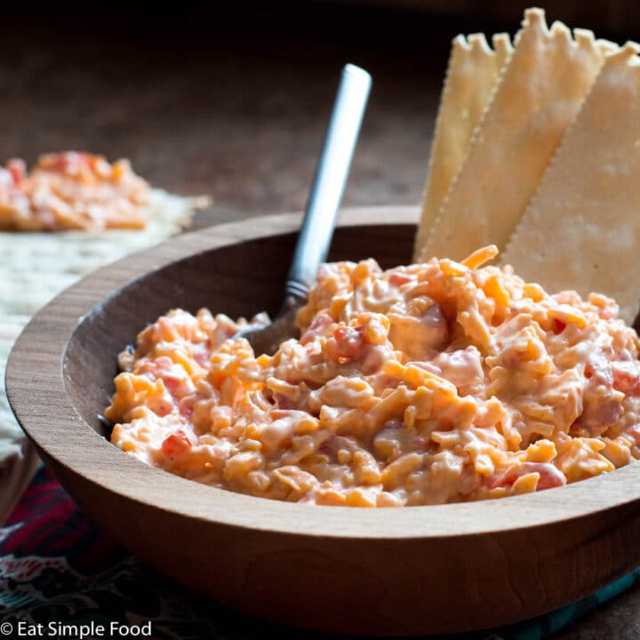 Wood bowl of pimento cheese with 3 crackers and a small spoon hanging out. Crackers on plate in background and one with pimento cheese on it.
