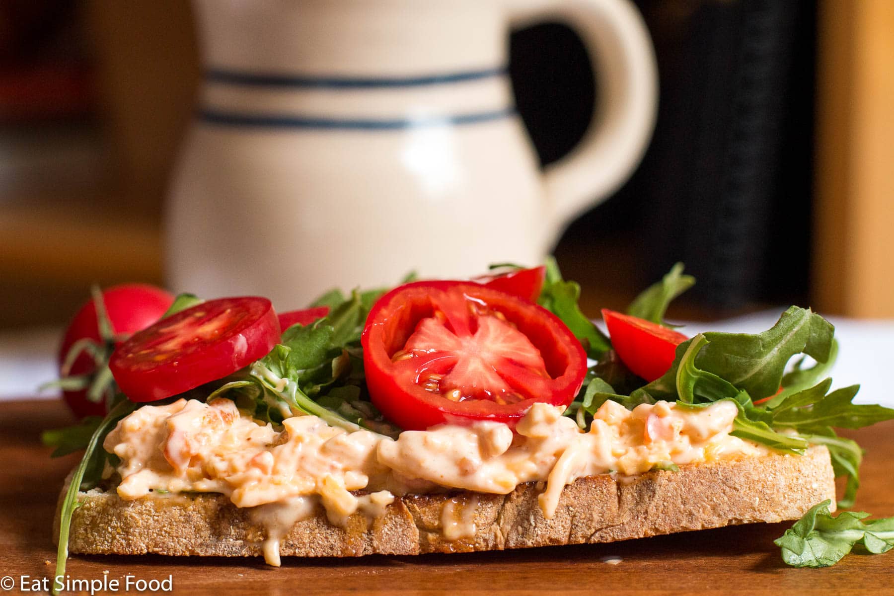 A piece of toast on a wood cutting board topped with pimento cheese, arugula, and sliced small tomatoes. 1 tomatoes in backdrop. side view.