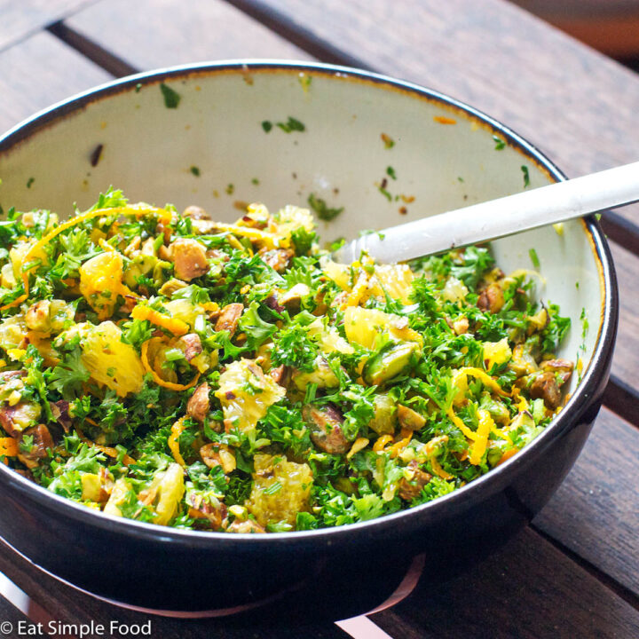 White Bowl with black outside filled with diced oranges, chopped parsley and pistachios with a silver spoon.