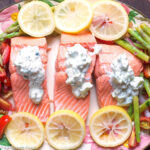 3 pieces of poached salmon on a platter surrounded by asparagus, quartered cherry tomatoes, walnuts, and lemon slices. Topped with Tzatziki Sauce