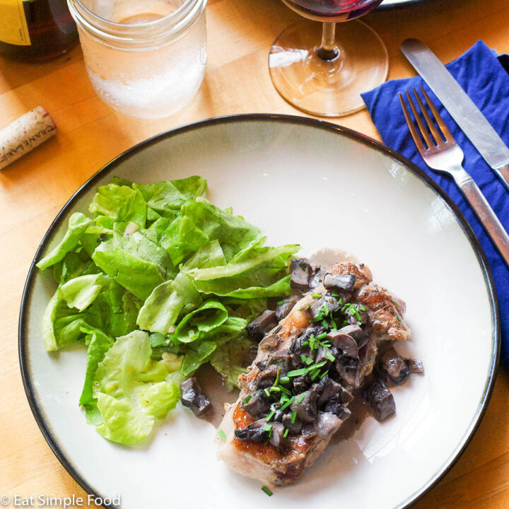 Pork Chop on a plate with a green lettuce salad. Chunky Portobello Mushrooms on top of chop. On a white plate with a red glass of wine.