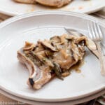 sdie View of a Pork Chop on a White plate smothered with chunky mushroom gravy. Fork and knife on the side of plate.