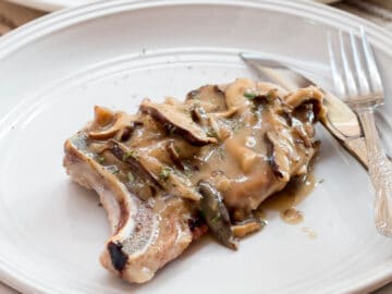 sdie View of a Pork Chop on a White plate smothered with chunky mushroom gravy. Fork and knife on the side of plate.