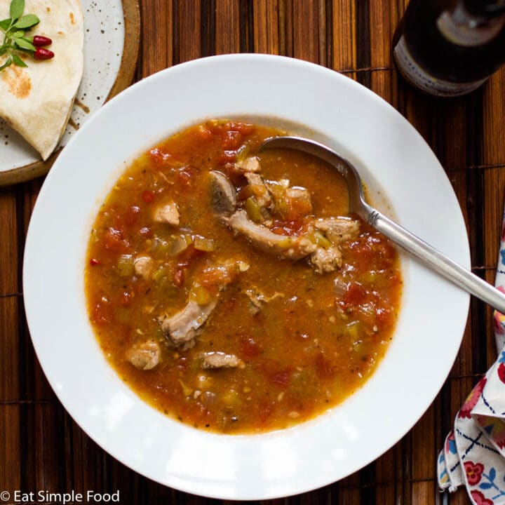 White shallow bowl filled with a red green sauce with pork and green chiles. Spoon hangiing out the bowl and tortilla and napkin on the side.