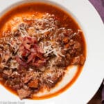 White shallow bowl of chunks of cooked pork shoulder and Italian Sausage in a red marinara sauce over grits. Garnished with Parmesan cheese and crispy chopped bacon.