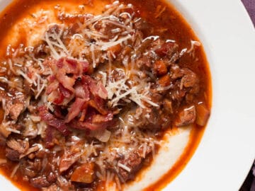 White shallow bowl of chunks of cooked pork shoulder and Italian Sausage in a red marinara sauce over grits. Garnished with Parmesan cheese and crispy chopped bacon.