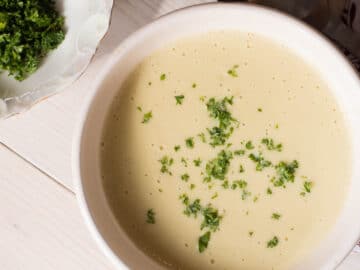 Creamy bowl of tan potato and leek soup on a white table with parsley garnish. top view.