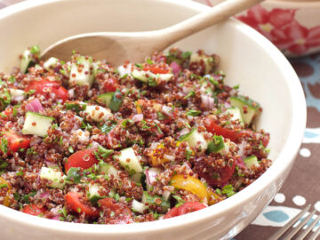 White bowl with wood spoon of red quinoa, diced tomatoes and cucumbers and red onions, and chopped parsley mixed together.