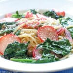 White deep plate of spaghetti with sautéed kale and thin sliced radishes. Topped with grated Parmesan cheese.