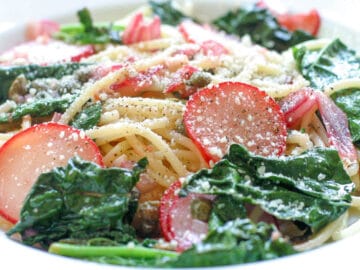 White deep plate of spaghetti with sautéed kale and thin sliced radishes. Topped with grated Parmesan cheese.