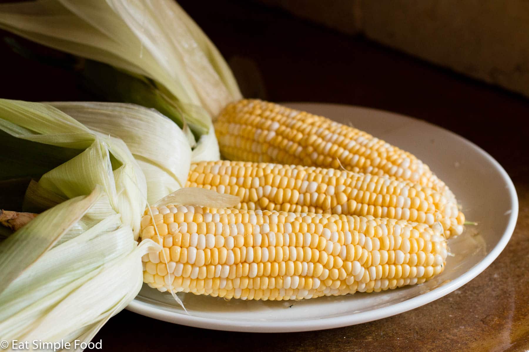 3 Corn on The cob on a white plate with husks pulled back and silks removed.