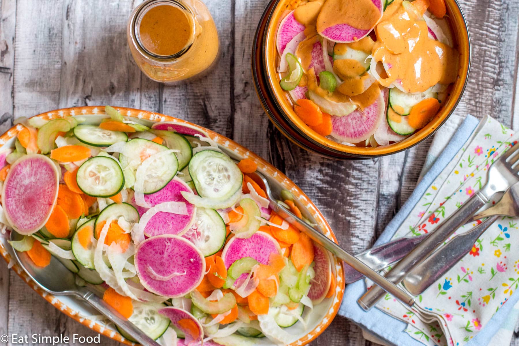 top view of oval shallow bowl and wooden small bowl filled with colorful sliced watermelon radishes, cucumber, fennel, carrots, and celery. Jar of red tomato dressing on the side.
