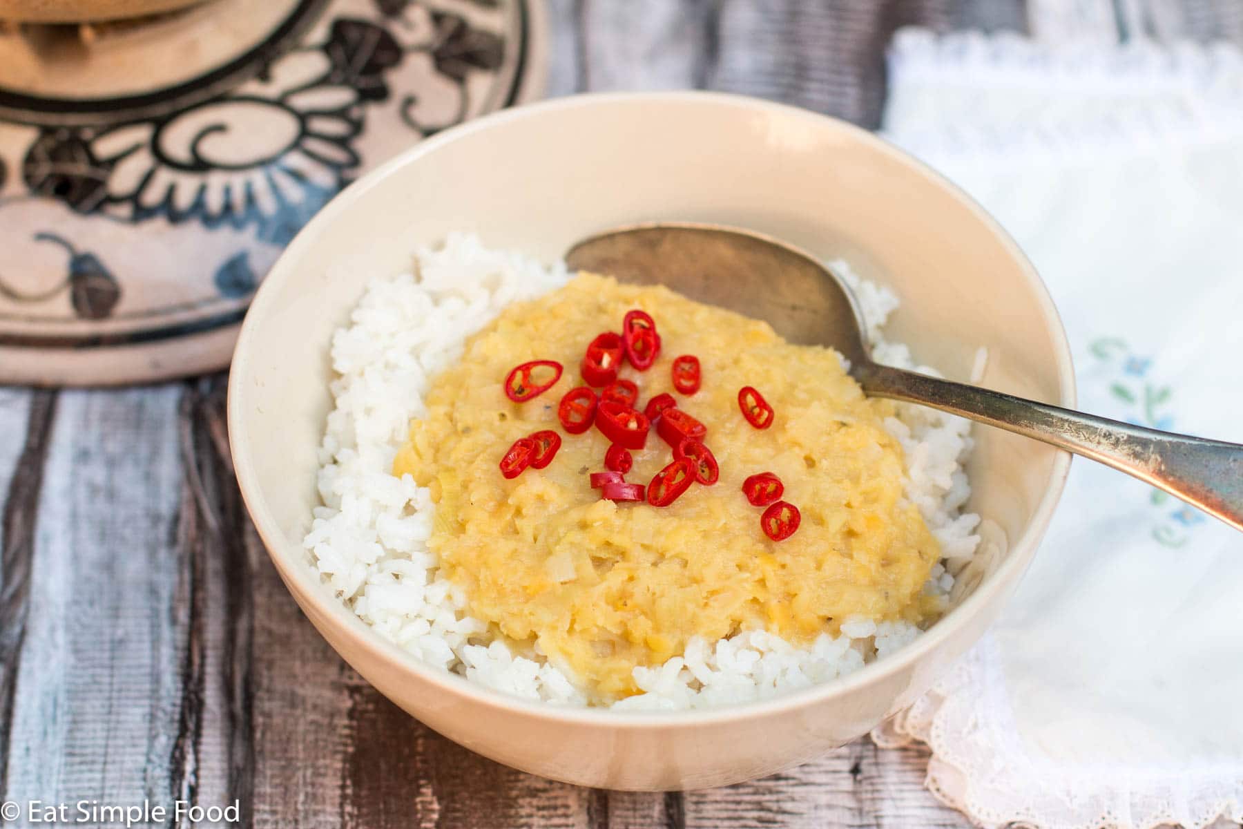 Light tan bowl of yellow cooked lentils over white rice with sliced red chili garnish.