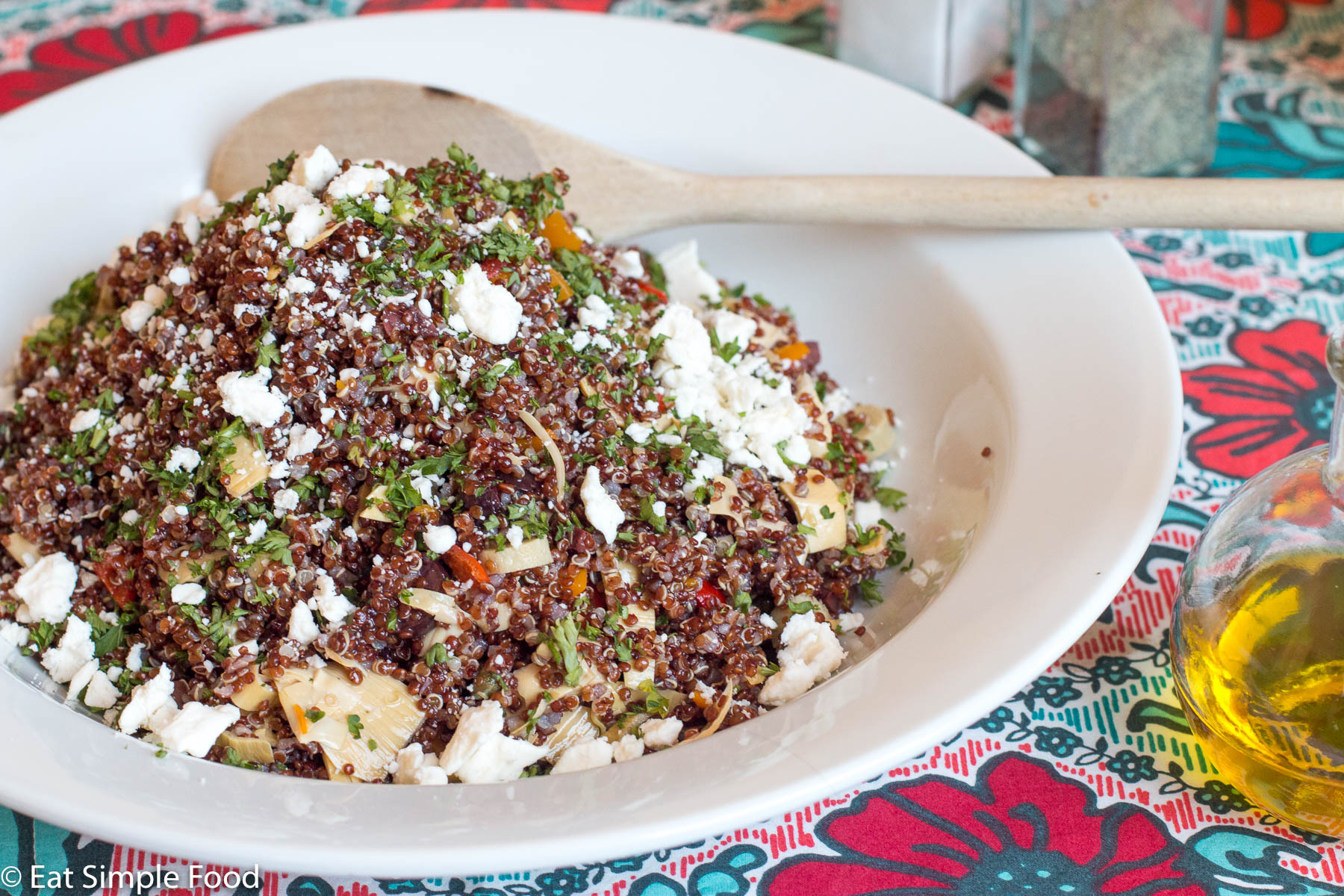 Top view of mound of red quinoa with diced red peppers, artichoke hearts, capers, and pepperoncinis topped with crumbled feta on a white plate.