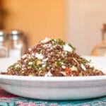 Side view of mound of red quinoa with diced red peppers, artichoke hearts, capers, and pepperoncinis topped with crumbled feta on a white plate.
