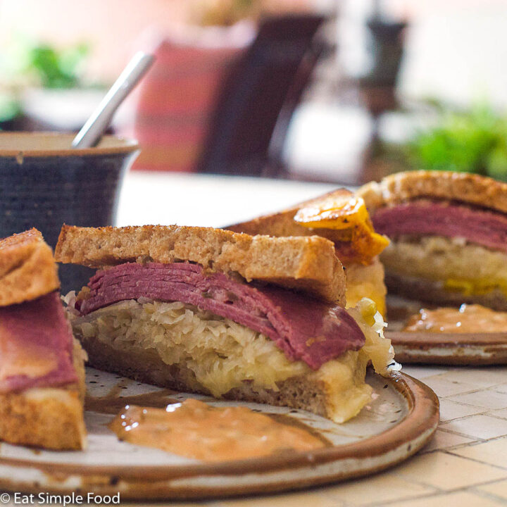 Halved Reuben Sandwich with pink corned beef, sauerkraut, melty swiss cheese with thousand islands dressing on a plate. Blue cup in background.