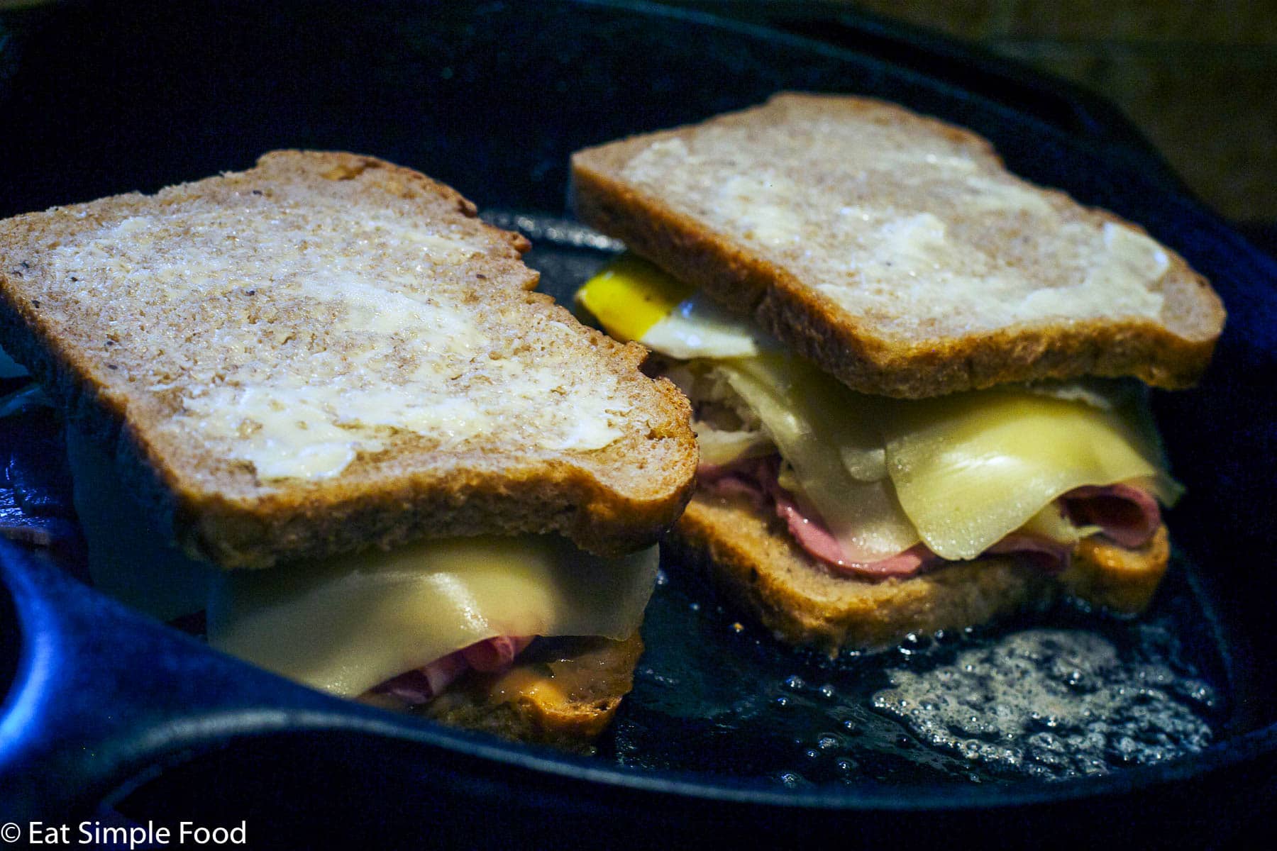 2 sandwiches being pan fried in a black cast iron skillet. The bread is buttered on the outside and white swiss cheese is melting.