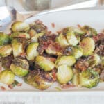 Roasted Brussels Sprouts with Crispy diced pancetta on a white platter with a large silver fork. side view.