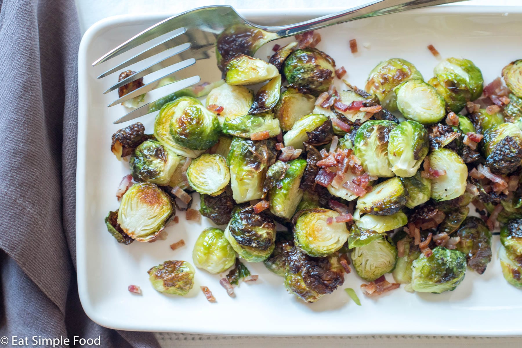 Roasted Brussels Sprouts with Crispy diced pancetta on a white platter with a large silver fork. top view.