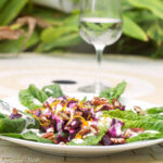 White plate of romaine lettuce, chunked beets and chopped pecans with a white dressing and orange zest garnish. Wine glass filled with water in the back. Outside table.