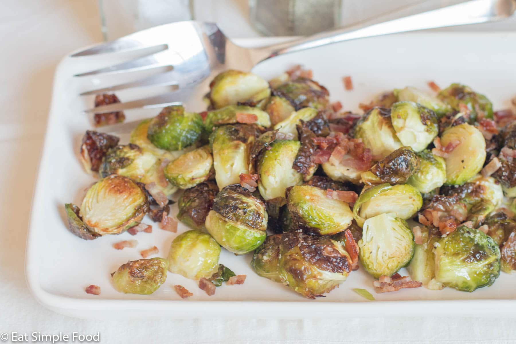 Roasted Brussels Sprouts with Crispy diced pancetta on a white platter with a large silver fork. side view.