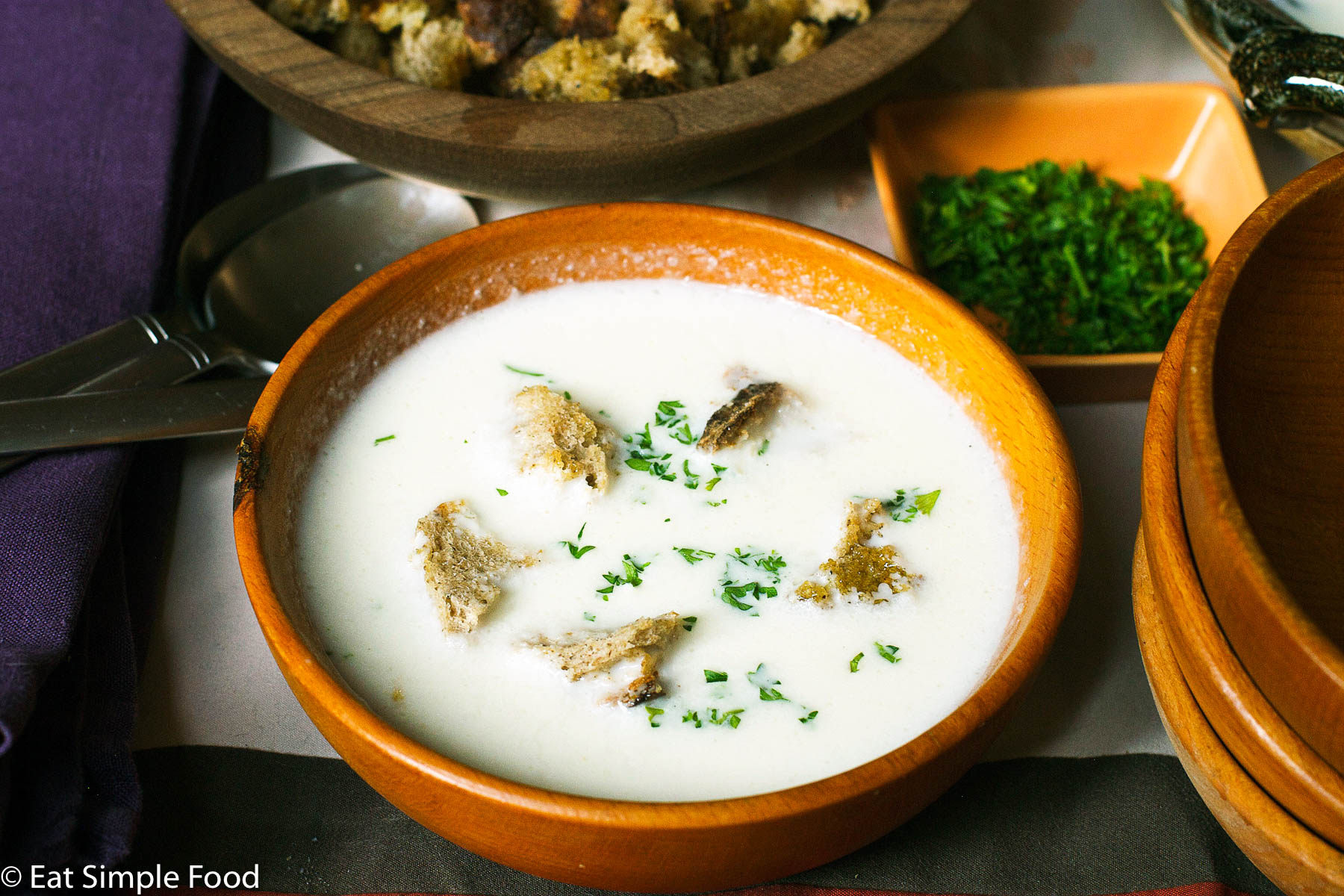 Side View wood bowl of white cauliflower soup with 5 croutons and a chopped parsley garnish