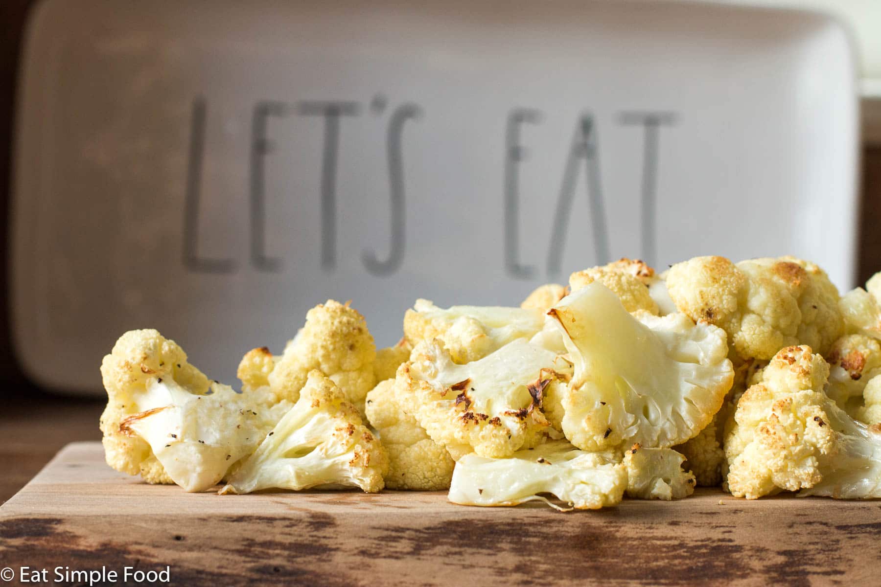 Cauliflower florets roasted brown on a wood cutting board with a white platter that says "let's eat" sitting behind it.