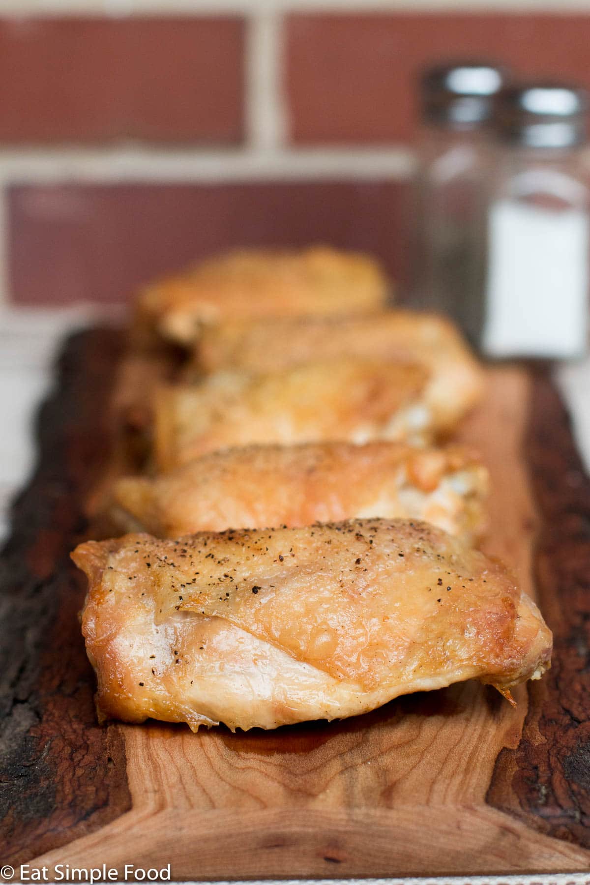 5 Oven Roasted bone in skin on chicken thighs on a wood cutting board with a salt shaker in the back.