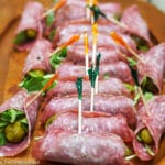Little Cornichon pickles wrapped with Arugula and rolled up in salami with colorful toothpicks holding them together. Ten of them on a wood plate.