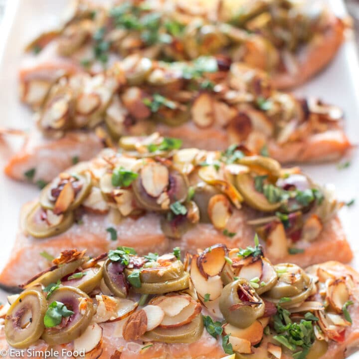 4 salmon fillets topped with sliced green olives, toasted sliced almonds, and parsley tapenade on a white rectangle plate with 4 lemon wedges.