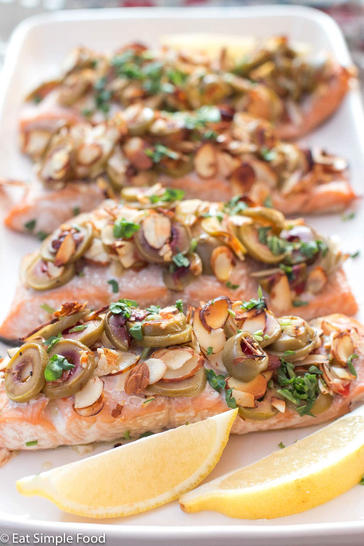 4 salmon fillets topped with sliced green olives, toasted sliced almonds, and parsley tapenade on a white rectangle plate with 4 lemon wedges.