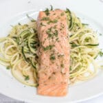 Salmon filet on a white plate on top of zucchini noodles with parsley and caper garnish. close up.