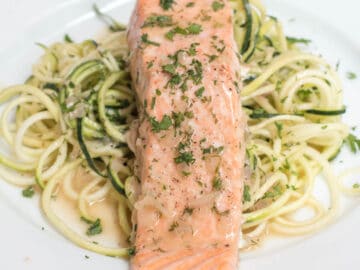 Salmon filet on a white plate on top of zucchini noodles with parsley and caper garnish. close up.