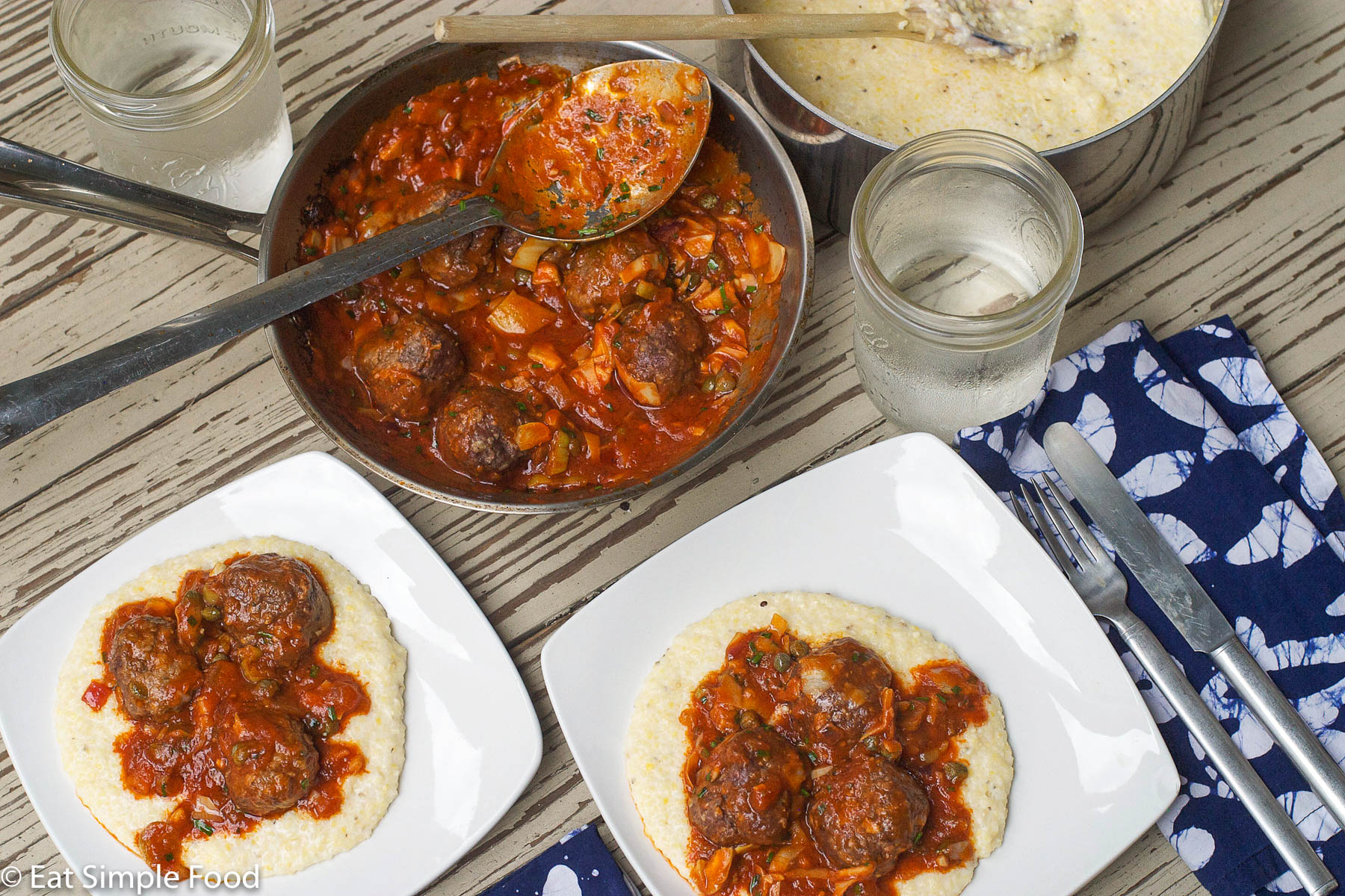 2 square plates with grits and meatballs in a red sauce. Glass of water and a shallow pan filled with meatballs and red sauce.