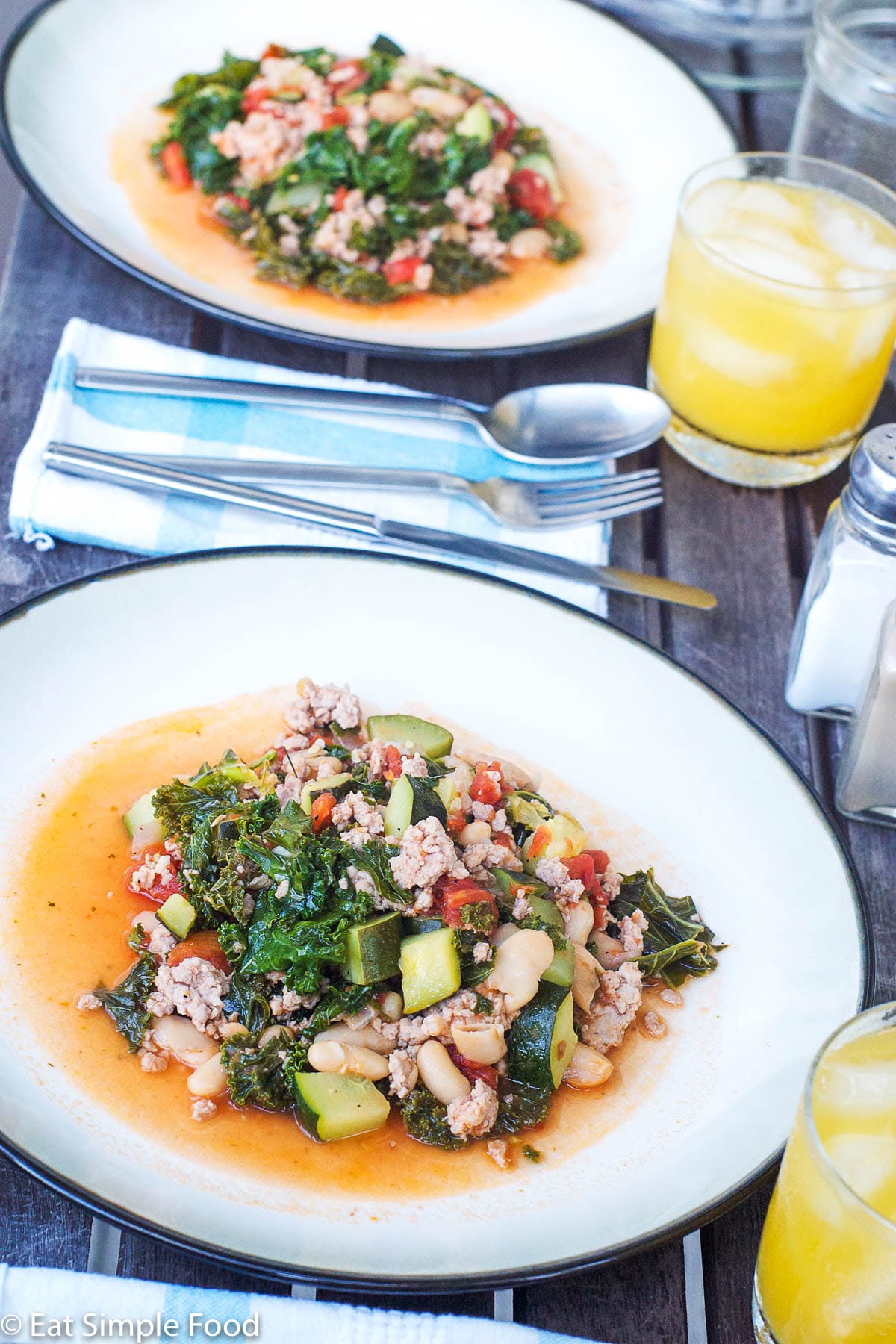 Ground sausage, chunks of kale, diced zucchini, tomatoes, and white beans on two white plates with silverware and orange drinks.