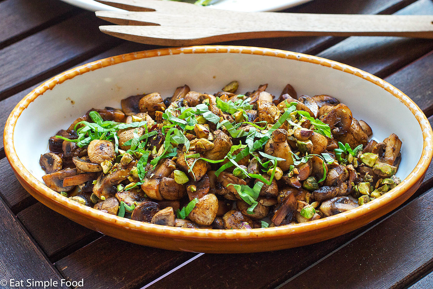 Cooked browned quartered mushrooms garnished with sliced basils and pistachios in a yellow baking dish.