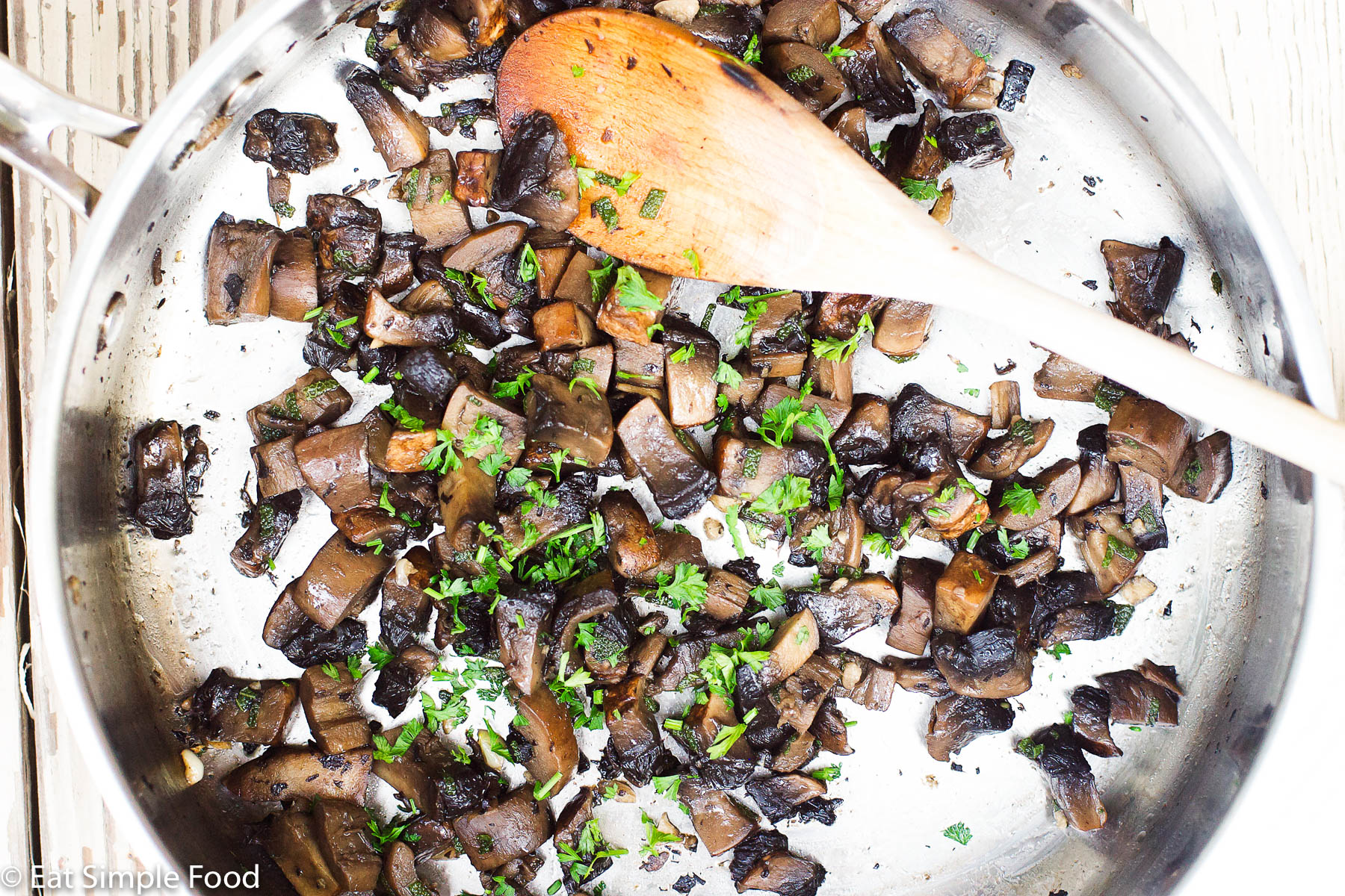 Stainless steel pan full of cooked Portobello Mushrooms with parsley garnish and a wood spoon sticking out of it.