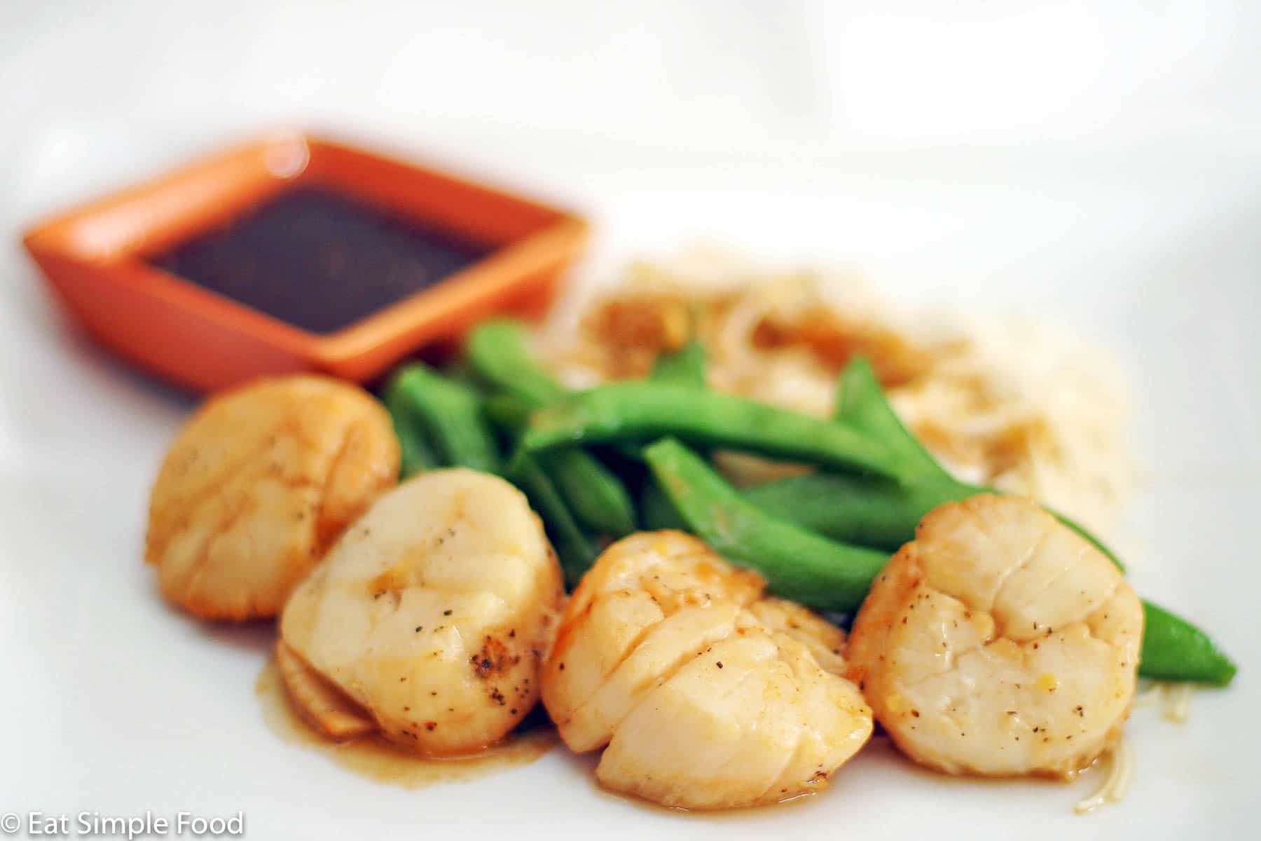 4 browned scallops in an orange sauce with sugar snap peas and rice and an orange small square container of soy sauce. Side view.