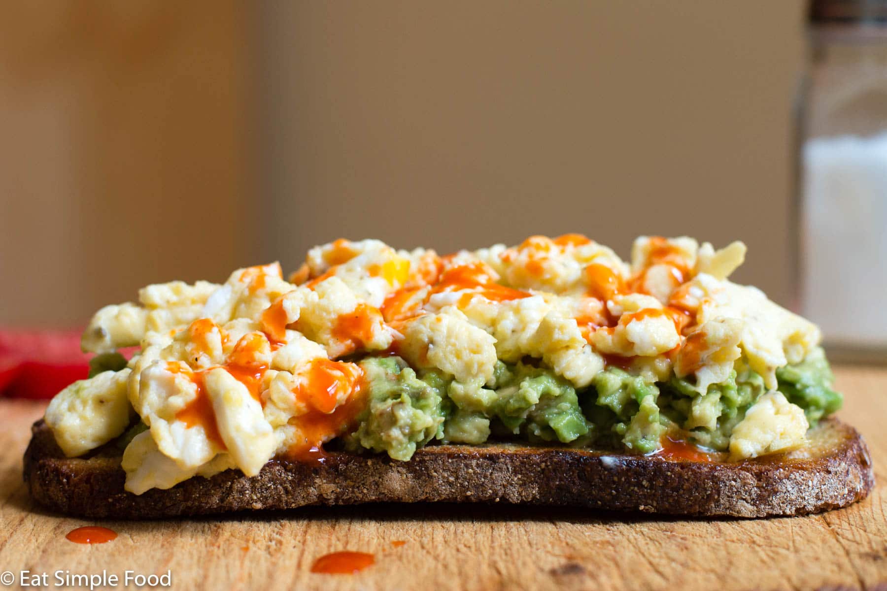 Side View Of Toast with Mashed Avocado, Scrambled Egg, and Hot Sauce On a Cutting Board