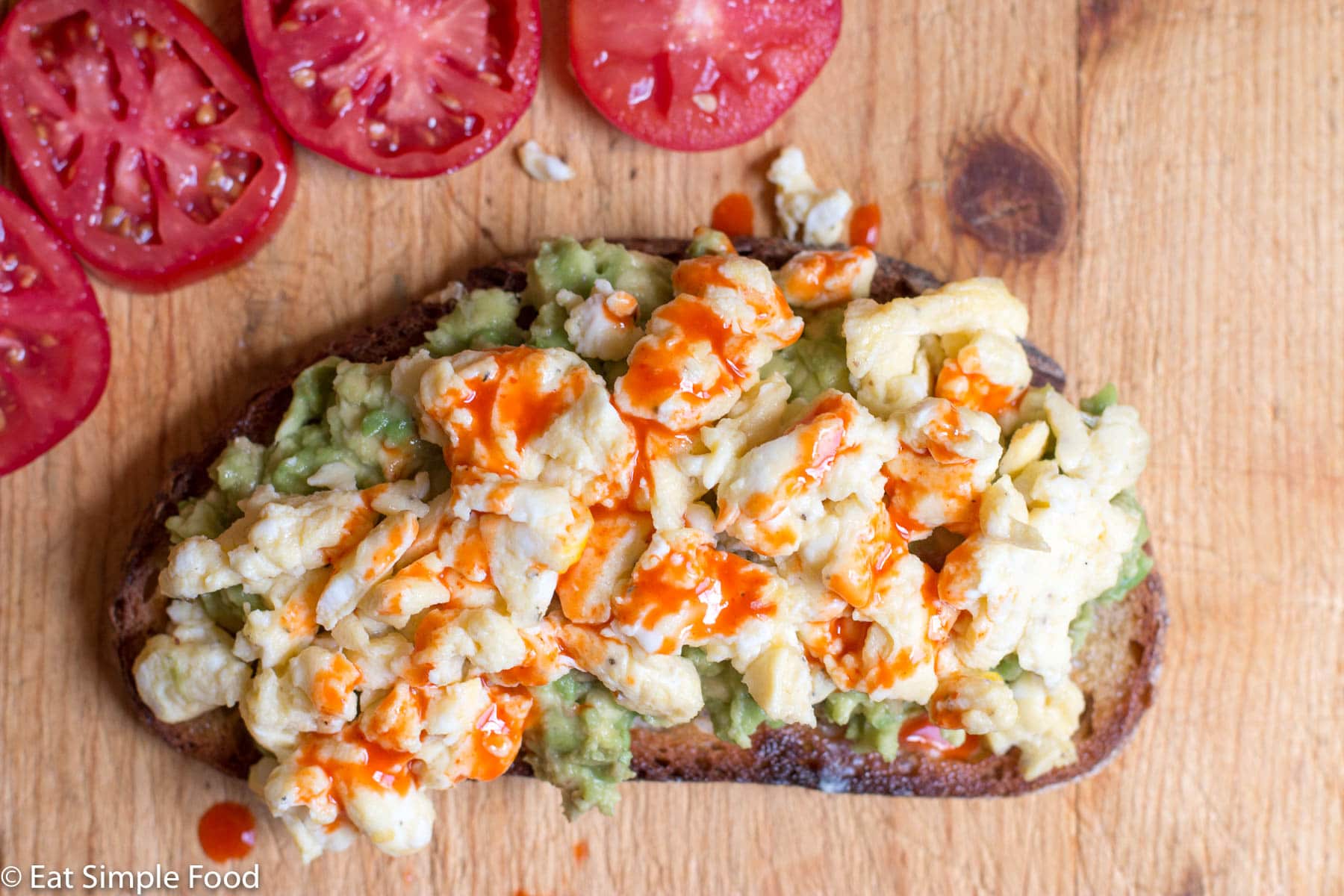 Top View Of Toast with Mashed Avocado, Scrambled Egg, and Hot Sauce On a Cutting Board with tomato Slices