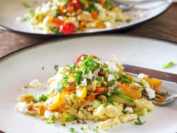 2 white plates of scrambled eggs topped with halved cherry tomatoes, bell peppers, parsley, and feta. Vertical view.