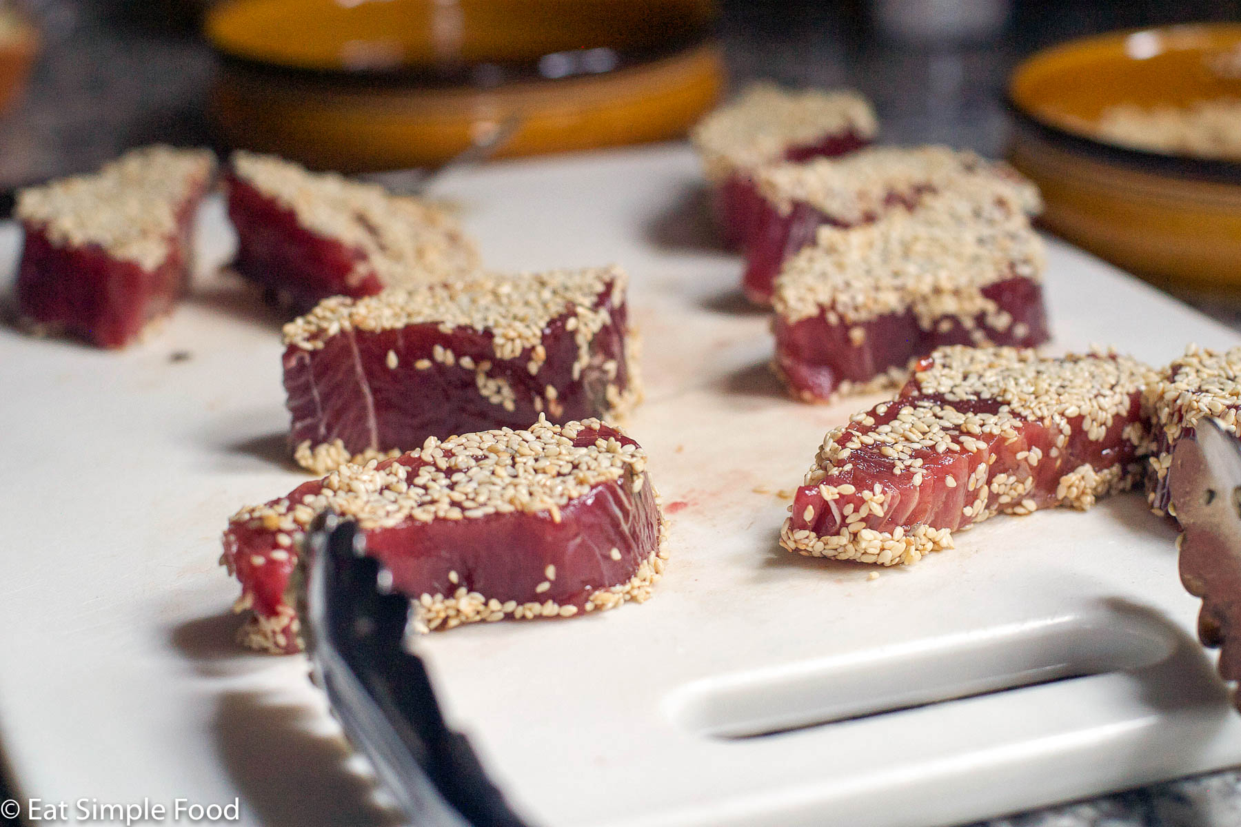 8 raw pieces of tuna coated in sesame seeds on a white cutting board with tongs