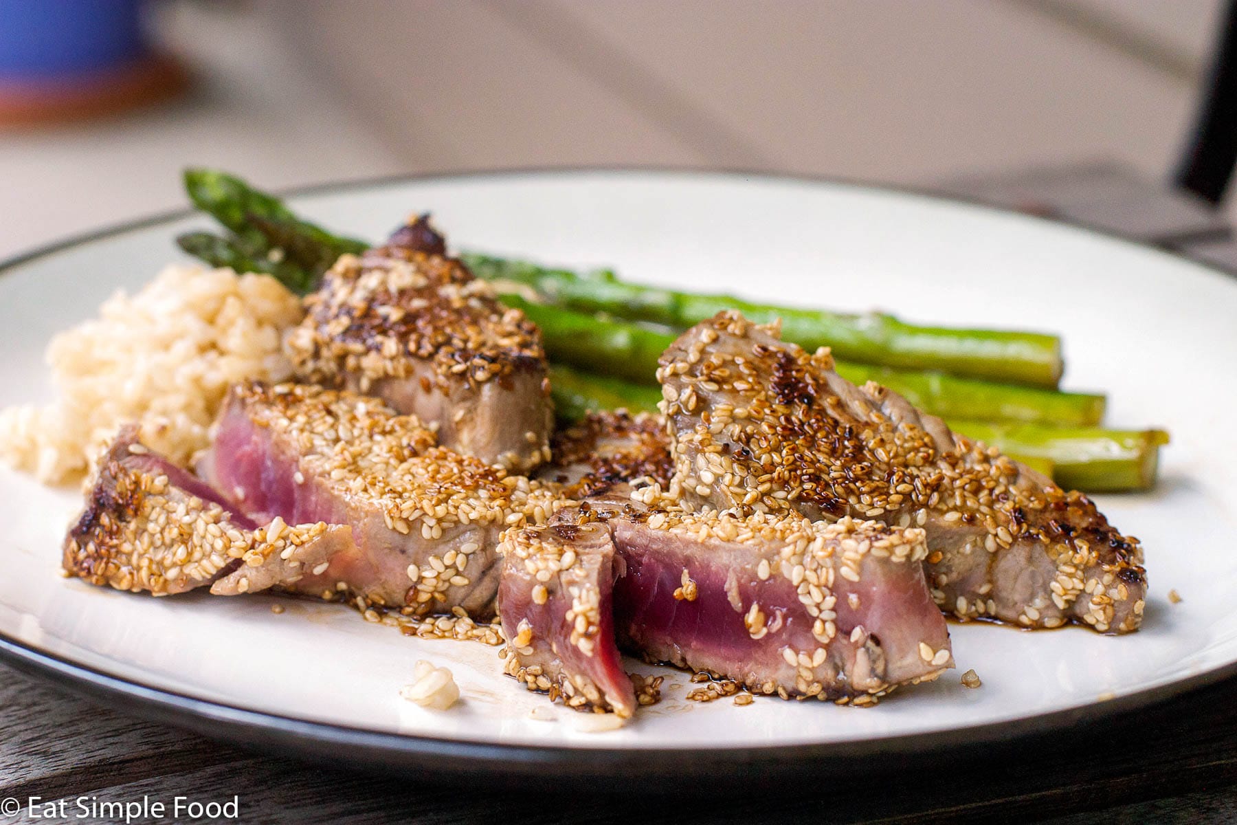 4 small pieces of tuna coated in sesame seeds and fried. One is cut open to expose the rare inside. On a plate with asparagus.