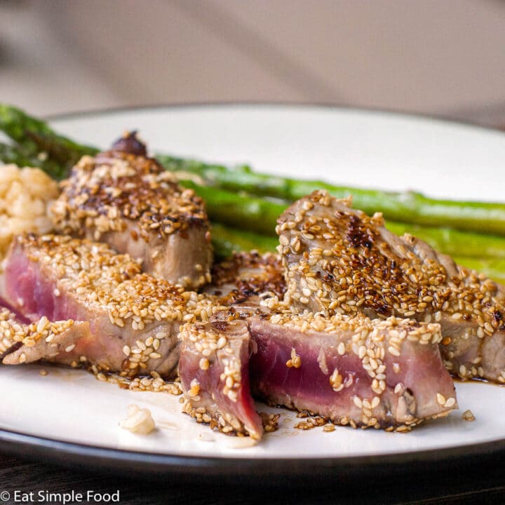 4 small pieces of tuna coated in sesame seeds and fried. One is cut open to expose the rare inside. On a plate with asparagus.