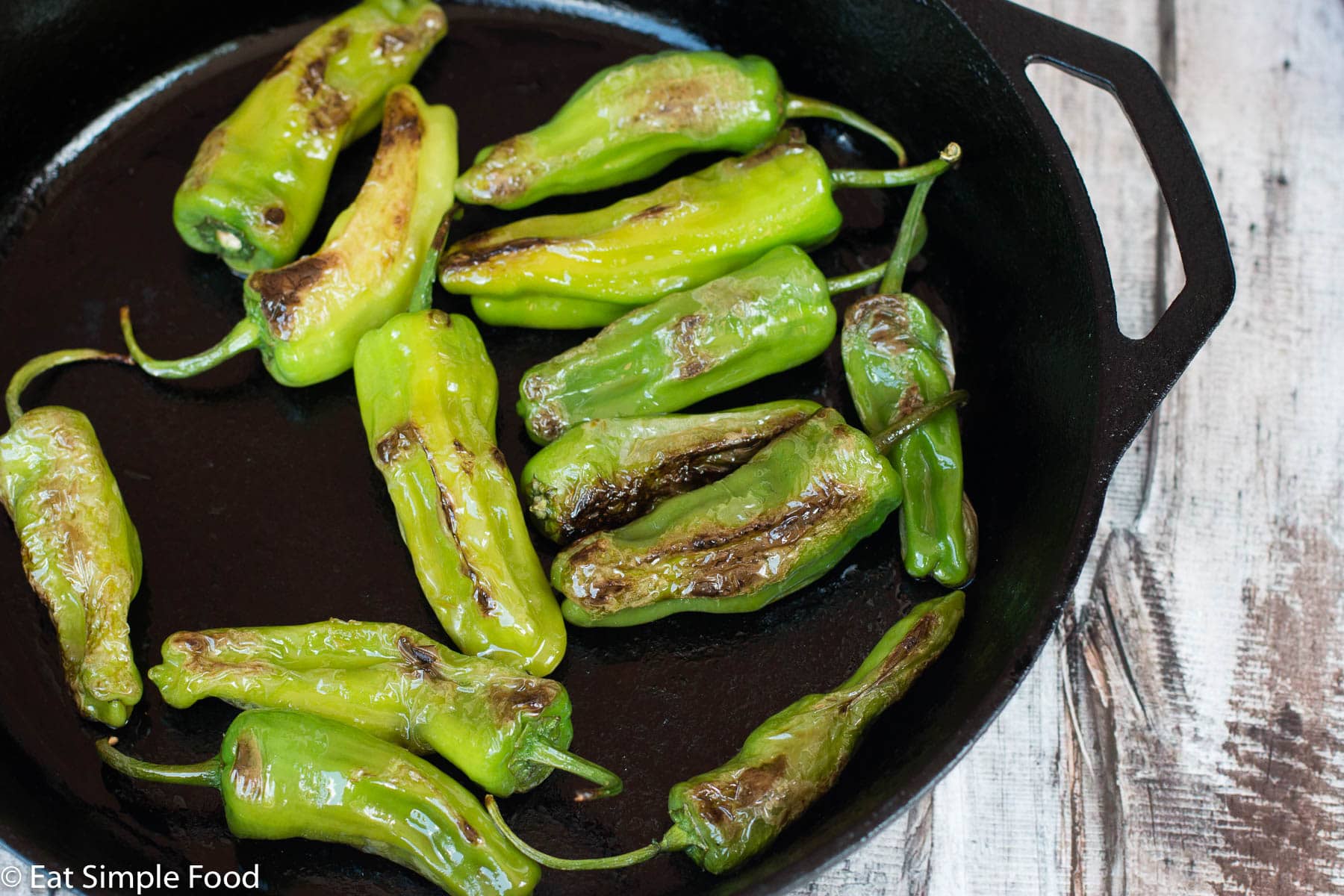 Green Shishito Peppers blistered with dark sear marks in a cast iron pan.