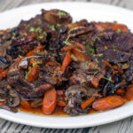 Beef Short Ribs and sliced carrots cooked in a rich gravy and plated and garnished with thyme. White plate. Side view.