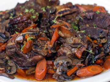 Beef Short Ribs and sliced carrots cooked in a rich gravy and plated and garnished with thyme. White plate. Side view.