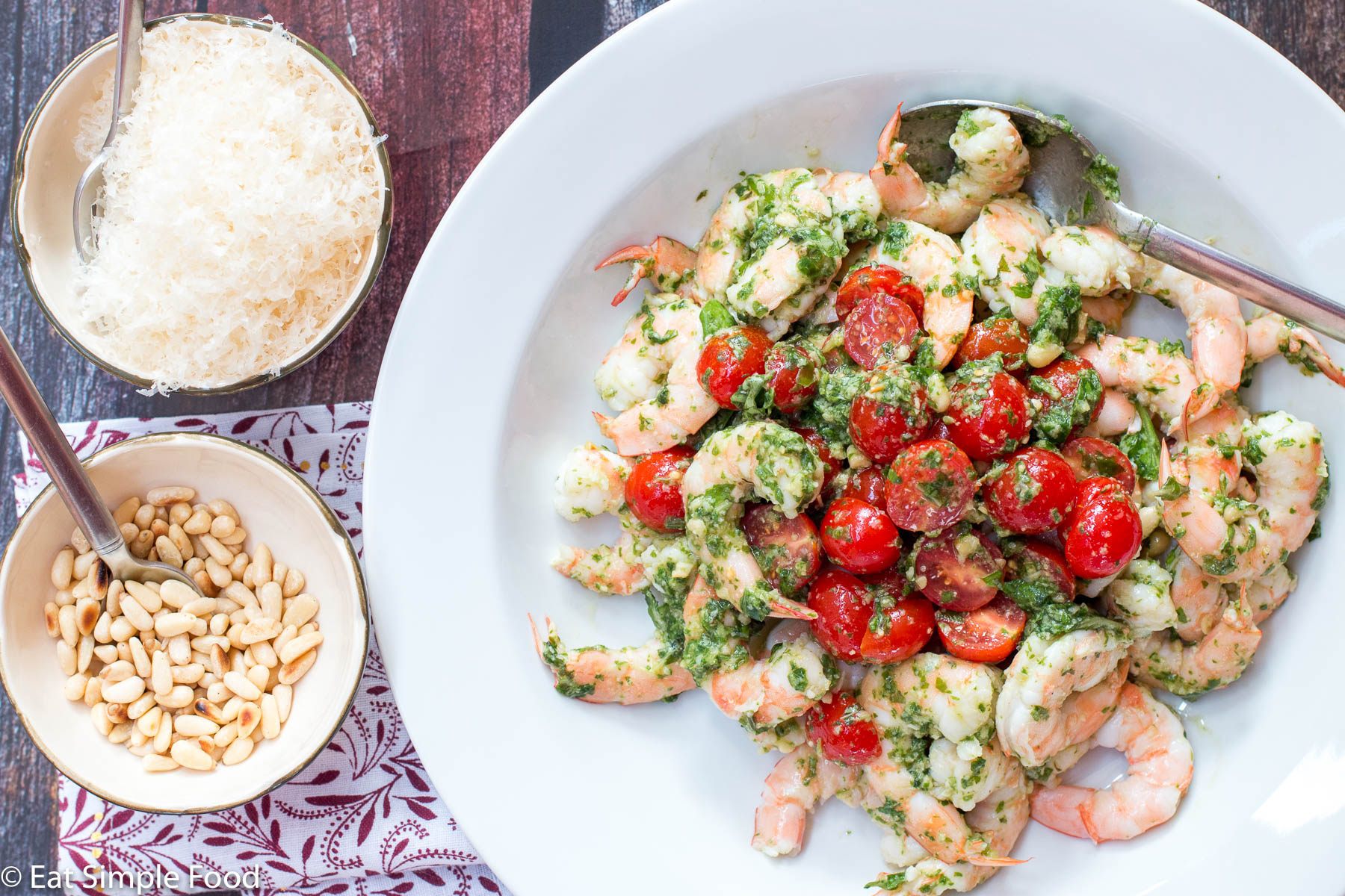 Cooked shrimp in a deep plate with halved cherry tomatoes and basil pesto. Pine nuts and Parmesan cheese in a bowl on the side.